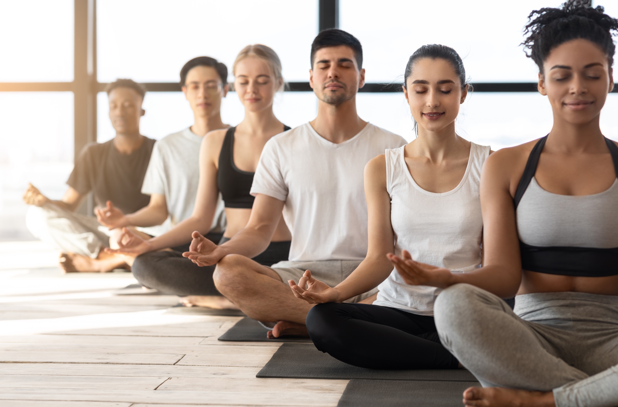 Group Meditation. Sporty Young Multiracial Men And Women Meditating Together During Yoga Class In Modern Light Studio, Sitting In A Row With Closed Eyes, Relaxing On Mats In Lotus Position, Free Space